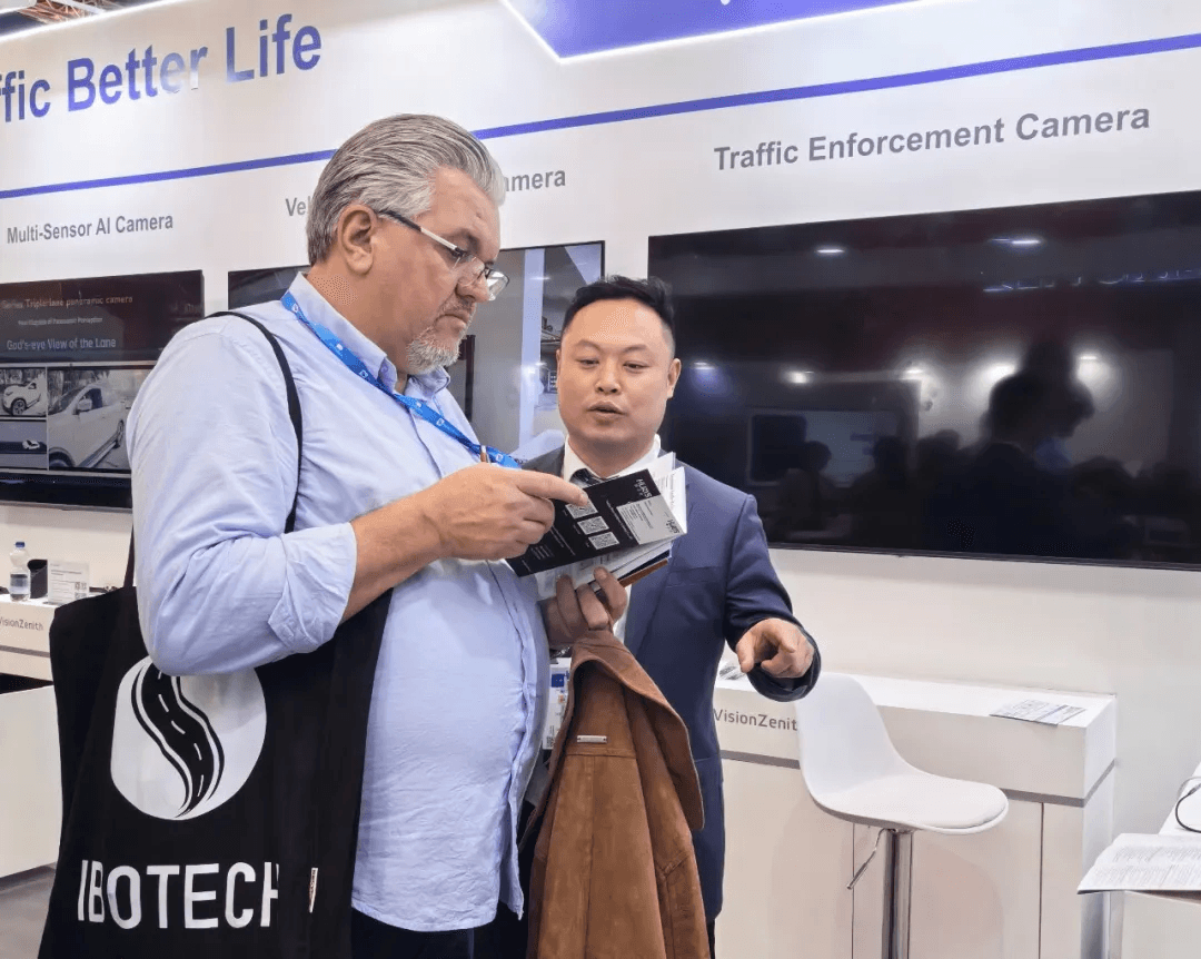Two men at a trade show booth discussing a brochure with a monitor in the background.